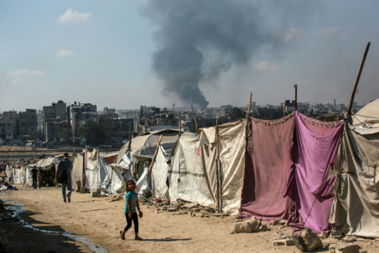 Des Palestiniens devant des tentes à Gaza-ville avec en arrière-plan une colonne de fumée s'élevant du centre de la bande de Gaza après un bombardement israélien, le 6 juillet 2025 ( AFP / Bashar TALEB )